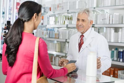 Smiling senior pharmacist looking at female making NFC payment for shampoo in pharmacy