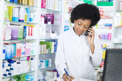 Young female chemist writing on clipboard while using cordless phone in pharmacy