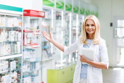 smiling female pharmacist holding digital tablet and showing medications in drugstore